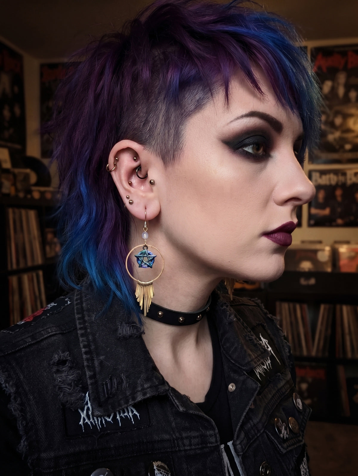 An ai generated model close-up side profile of a person with a purple and blue mohawk hairstyle, wearing a gold circular earring with a blue star and feather charm, a black choker, and a distressed black denim jacket in a record store setting.