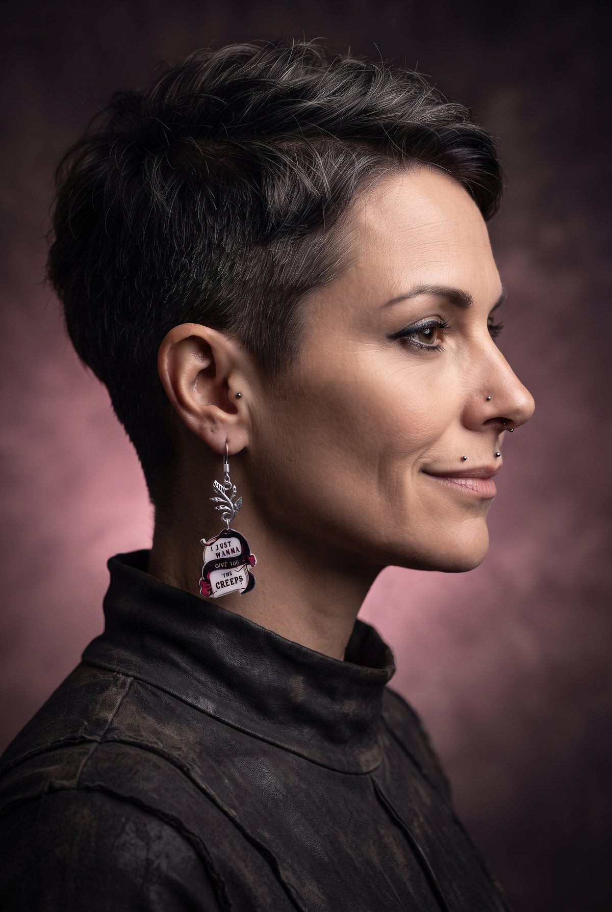 Wanna Give You The Creeps Earrings by Circle of Heather worn by a model with a short dark pixie cut, printed acrylic charm with a black snake and pink flowers hanging from a silver-tone leaf branch, against a black studio background.