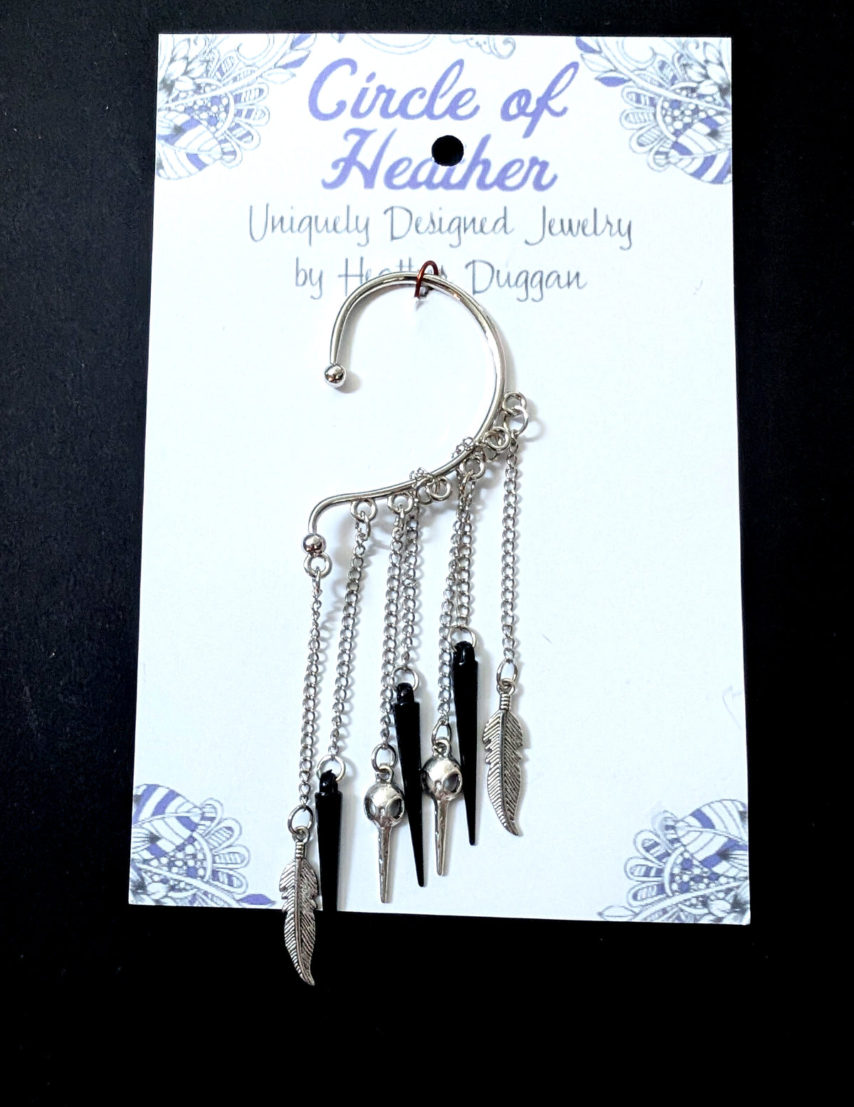 A silver bird skull ear cuff attached to a white backing card featuring a decorative purple mandala pattern and text that reads Circle of Heather, Uniquely Designed Jewelry by Heather Duggan.