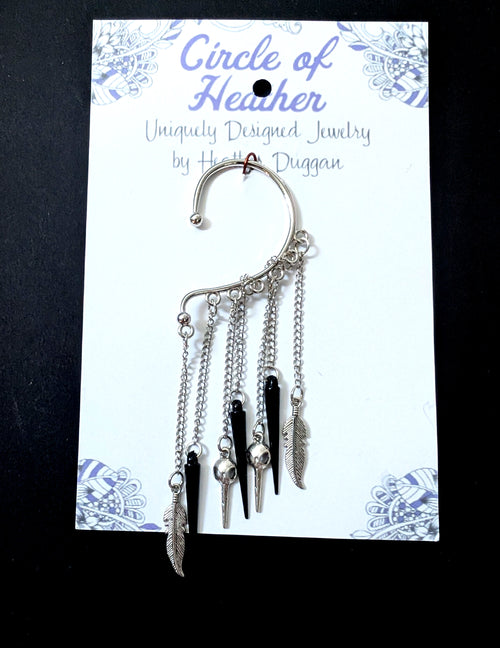 A silver bird skull ear cuff attached to a white backing card featuring a decorative purple mandala pattern and text that reads Circle of Heather, Uniquely Designed Jewelry by Heather Duggan.