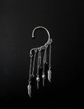 Silver ear cuff jewelry piece against a solid black background, featuring a curved upper cuff, hanging chains, feather charms, and small skull pendants.