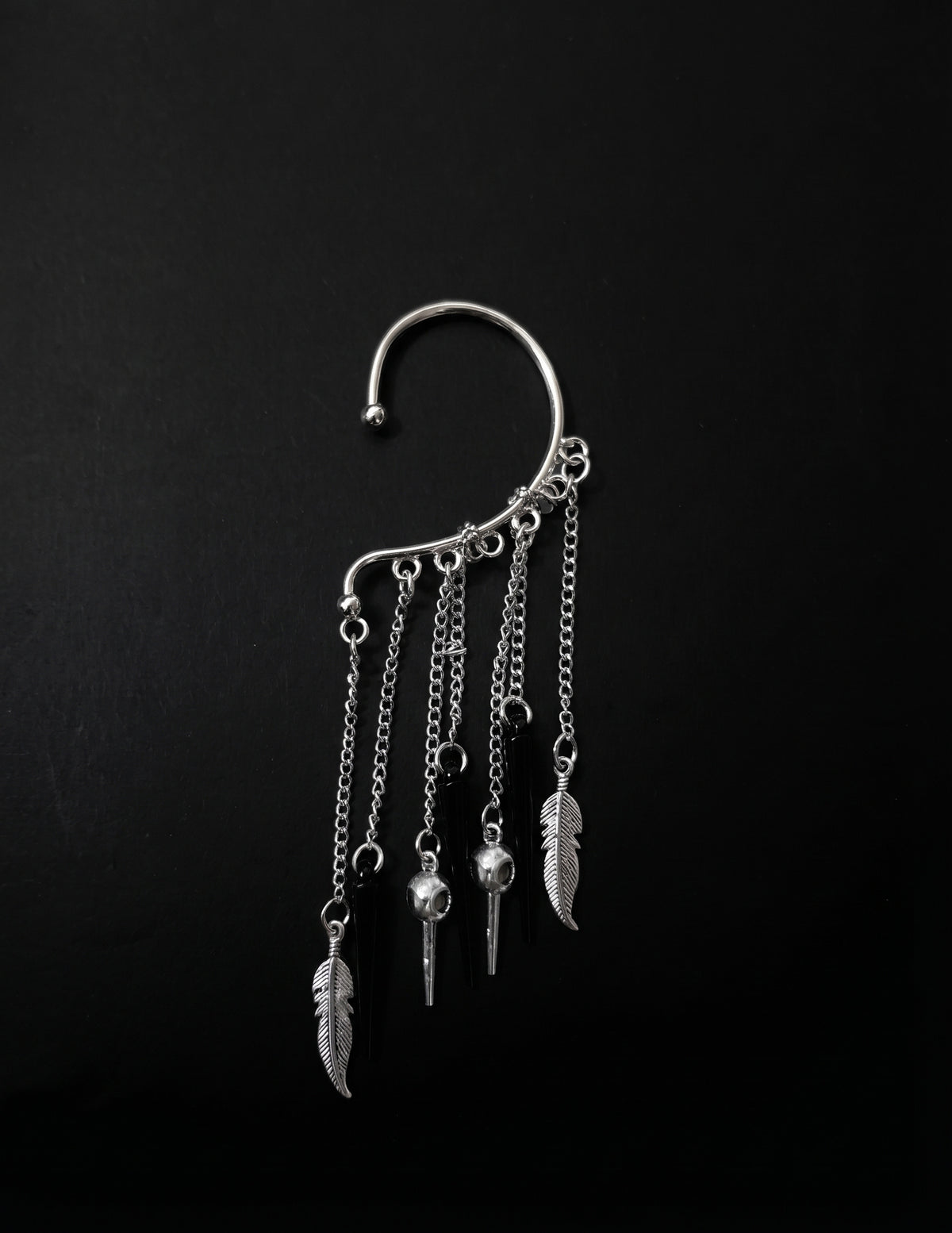 Silver ear cuff jewelry piece against a solid black background, featuring a curved upper cuff, hanging chains, feather charms, and small skull pendants.
