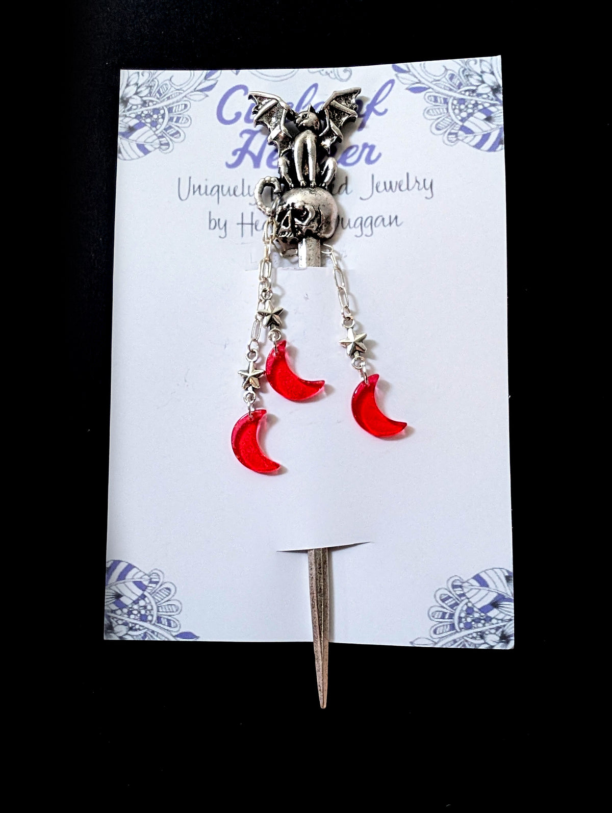 The silver winged creature and skull hair pick attached to a white backing card featuring a decorative purple mandala pattern and text that reads "Circle of Heather, Uniquely Designed Jewelry by Heather Duggan."