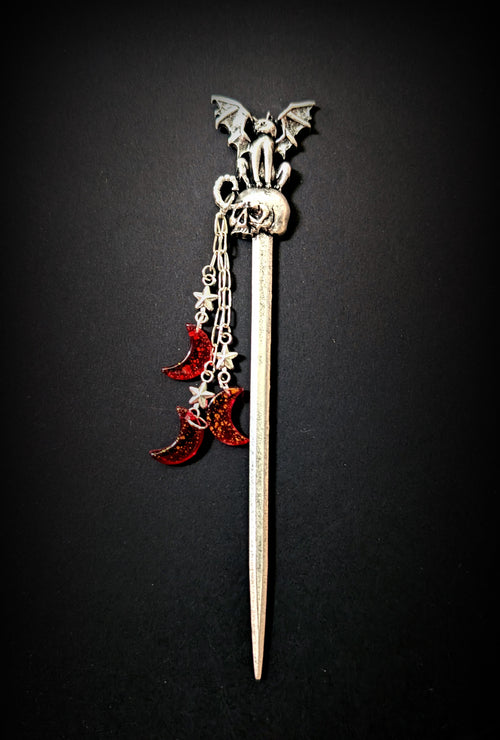 Isolated product shot of the silver hair pick against a black background, featuring a tapered shaft, a skull topper with a perched winged cat creature, and three dangling chains with silver stars and red glitter resin crescent moons.