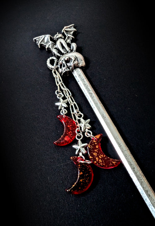 Close-up product shot against a black background, highlighting the detailed winged creature perched on the skull topper and the layered copper-glitter red resin crescent moon charms.