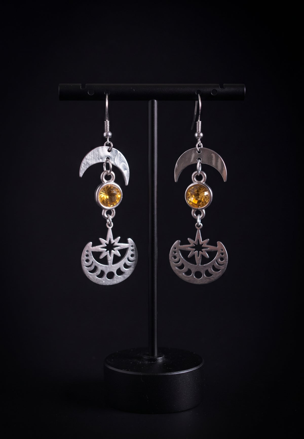 Celestial Sparkling Earrings by Circle of Heather, crescent moon tops with golden stones and cut moon phase discs, hanging on a black T-bar jewelry stand against a black background.