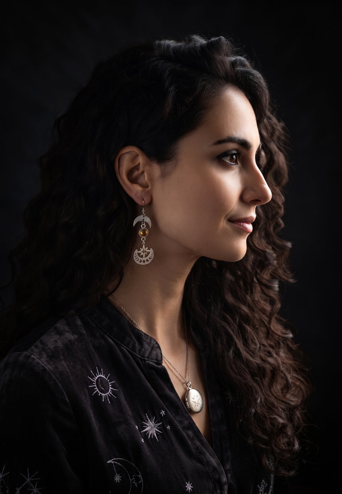 Celestial Sparkling Earrings by Circle of Heather worn by a model with long dark curly hair, silver crescent moon above a golden stone and a cut star and moon phase disc, shown with a black velvet top embroidered with suns, moons and stars.