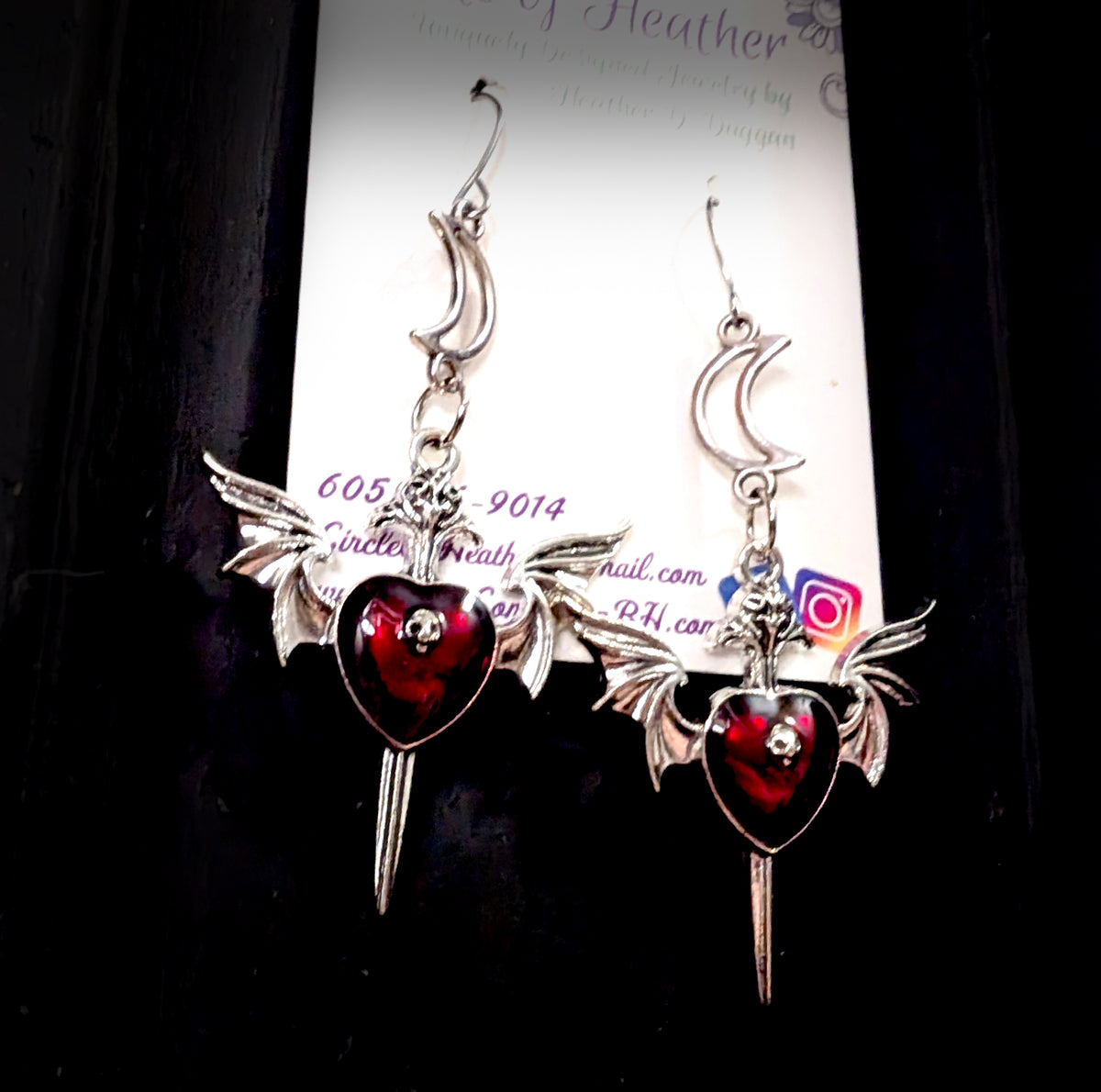 Close-up of Love Soars Earrings by Circle of Heather on packaging card, detailed view of the crescent moon, winged heart, and dagger charm design in red enamel
