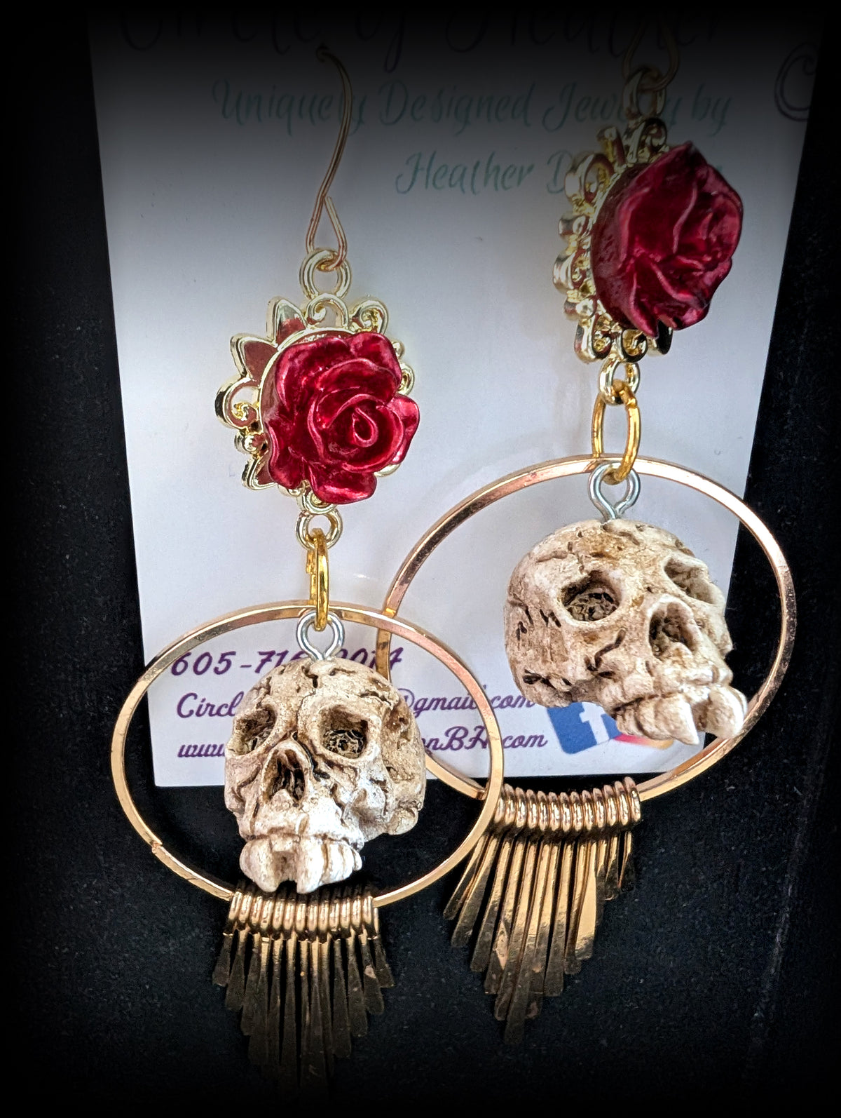 A pair of gothic dangle earrings featuring red roses, skull charms, and gold-tone circular hoops with fringe, displayed on a white card against a black background.