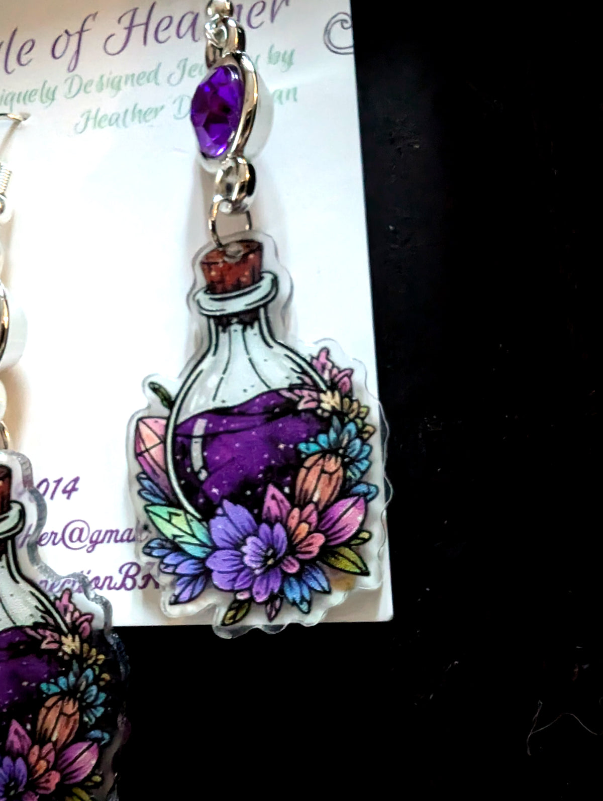 A close-up of a pair of acrylic earrings featuring a colorful potion bottle design with purple liquid, flowers, and a purple gemstone, displayed on a branded card against a black background.