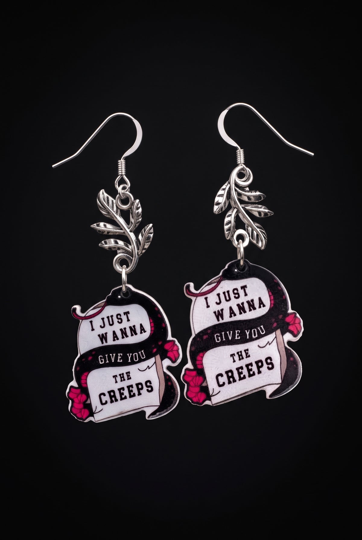Wanna Give You The Creeps Earrings by Circle of Heather, full view of both printed acrylic charms reading "I just wanna give you the creeps" with silver-tone leaf branches and fishhook earwires, on a plain black background.