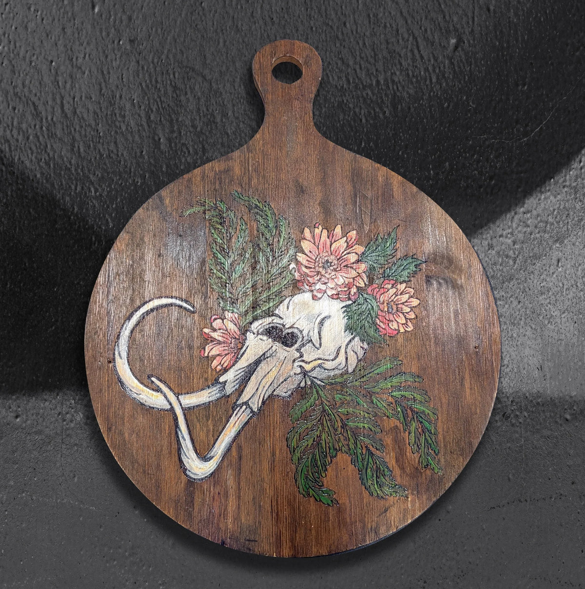 Dystopian Cyclopean by CamiThunderInk, an original pyrography and paint piece on a reclaimed round wood paddle showing a one eyed skull with tusks among pink chrysanthemums and green cedar