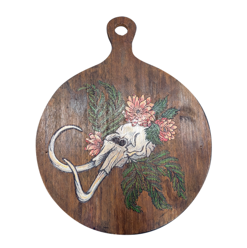 Another view of Dystopian Cyclopean by CamiThunderInk, an original pyrography and paint piece on a reclaimed round wood paddle showing a one eyed skull with tusks among pink chrysanthemums and green cedar