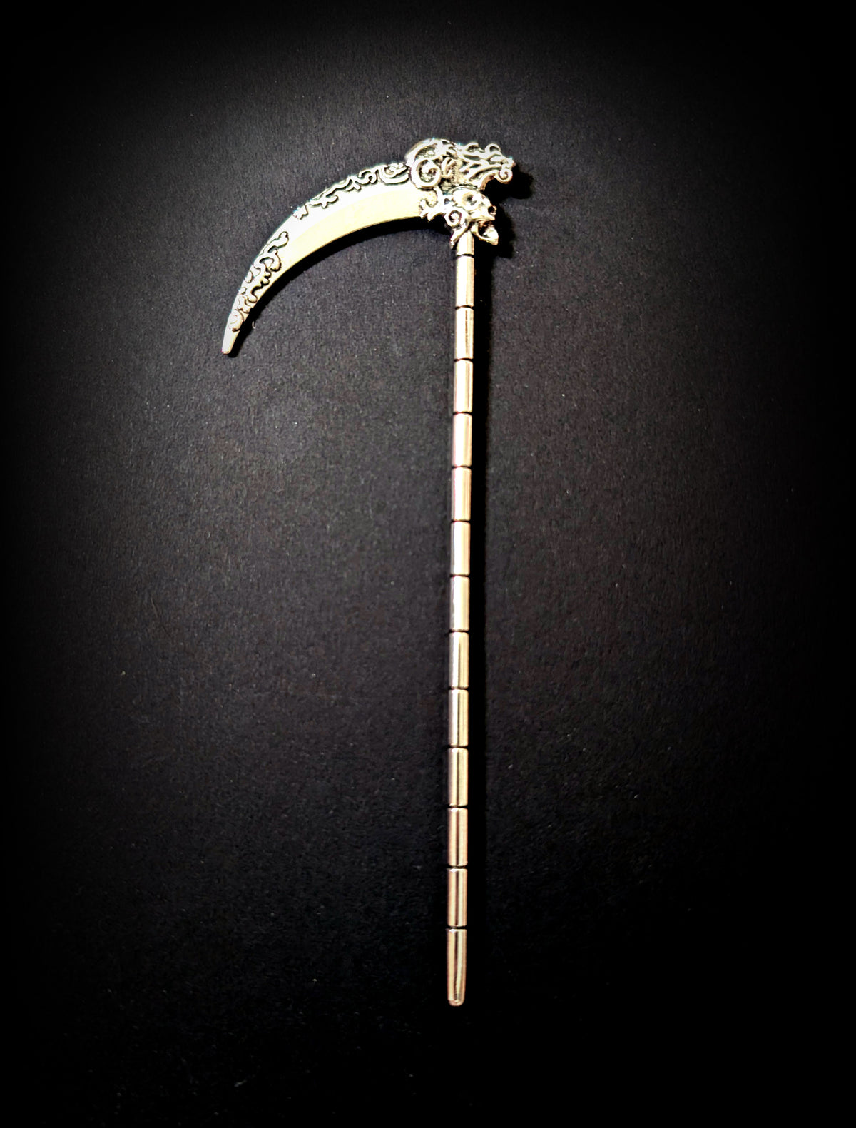 Isolated product shot of the silver scythe hair pick against a pure black background, showing the full length of the ribbed shaft, the skull joint, and the filigree-etched blade.