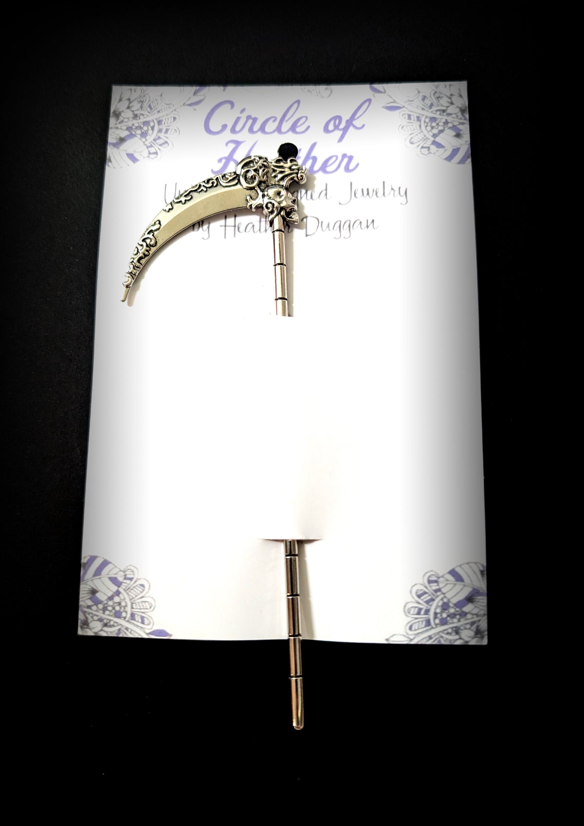 The silver scythe hair pick attached to a white backing card featuring a decorative purple mandala pattern and text that reads "Circle of Heather, Uniquely Designed Jewelry by Heather Duggan."