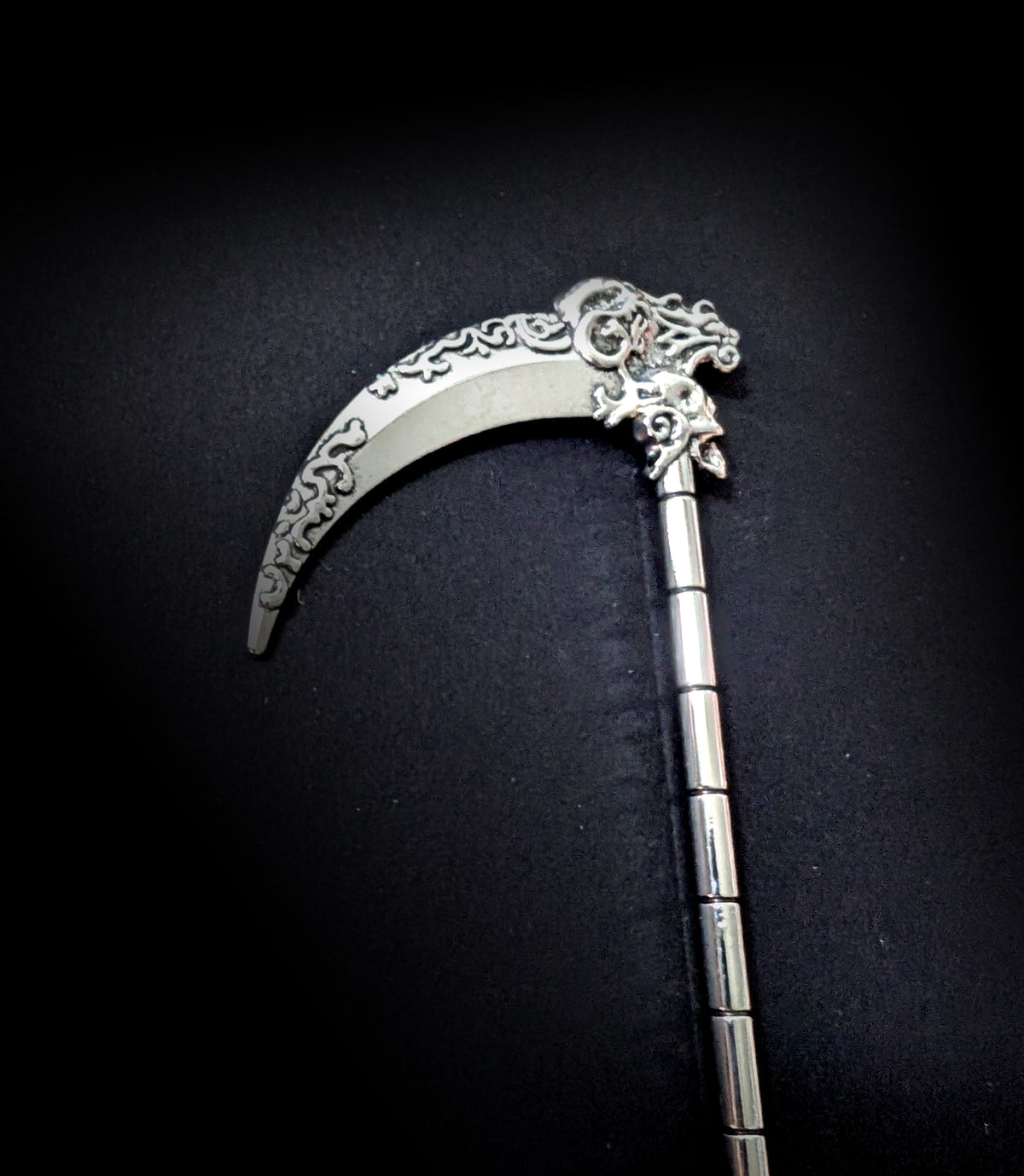 Close-up view of the silver scythe hair pick against a dark background, highlighting the detailed scrollwork on the blade curve and the small carved skull at the top of the ribbed shaft.