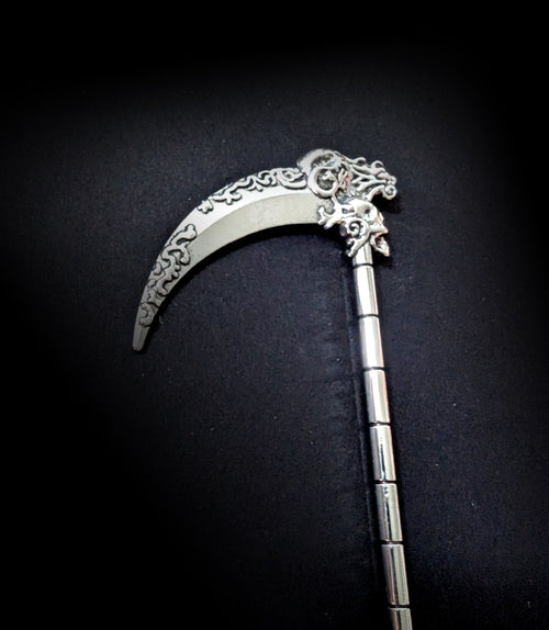 Close-up view of the silver scythe hair pick against a dark background, highlighting the detailed scrollwork on the blade curve and the small carved skull at the top of the ribbed shaft.