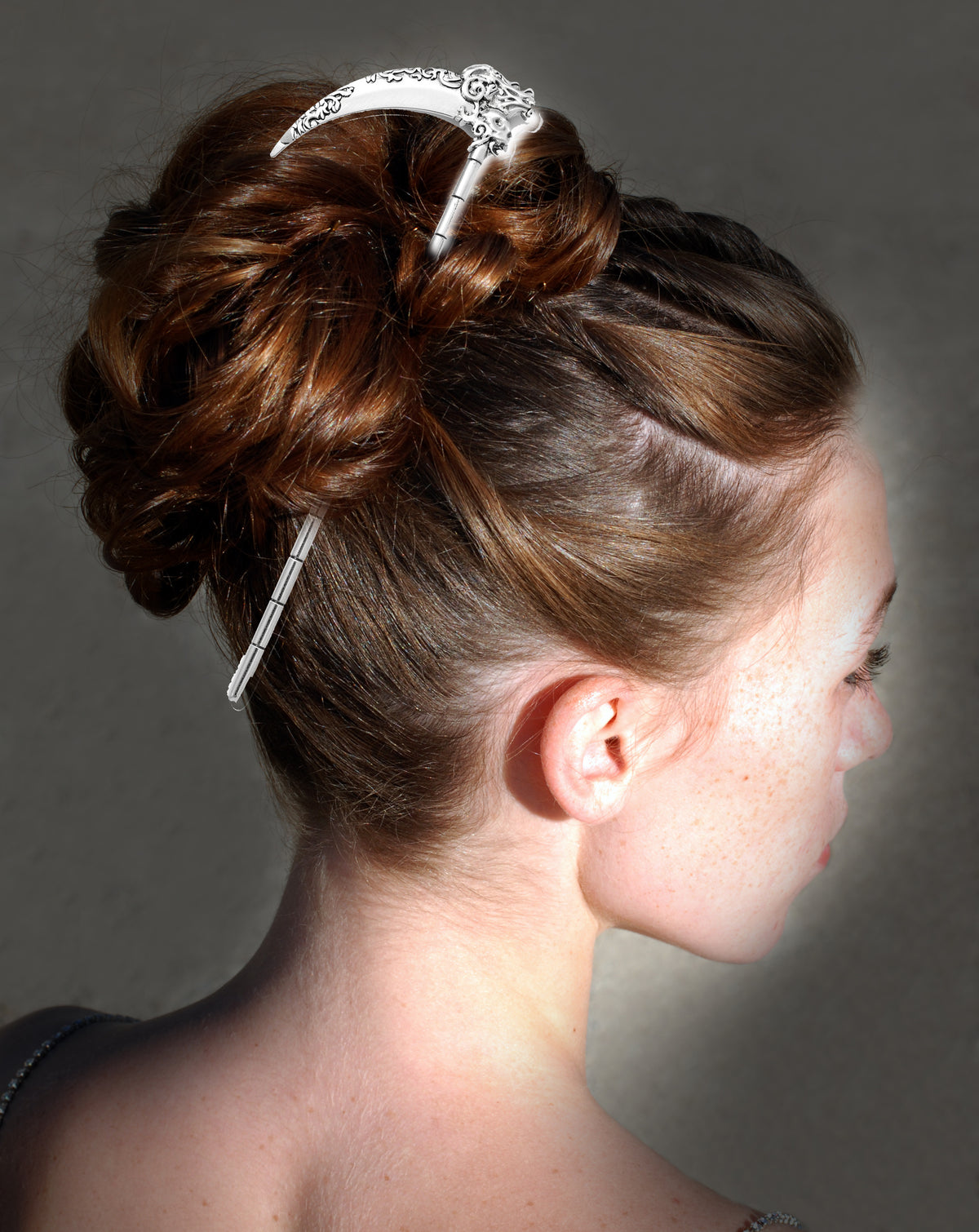 Side profile of a model with dark reddish-brown hair styled in a large bun secured by the silver scythe hair pick, set against a dark textured background.