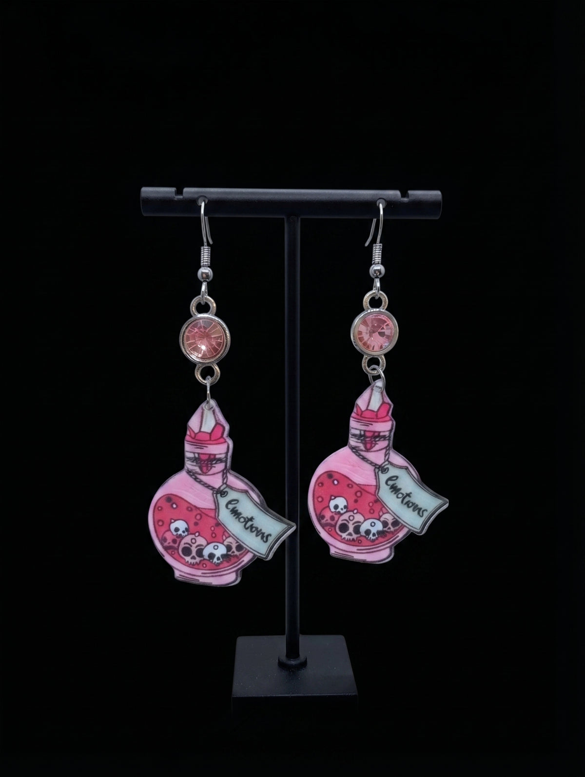 A pair of dangling earrings displayed on a black stand against a black background. Each earring features a pink potion bottle charm with a cartoon character and the word 'Enchant!' on a silver-toned hook with a round pink bead.