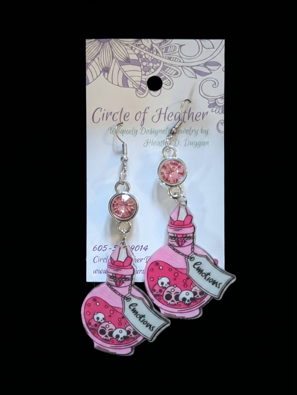 Pink and silver dangle earrings featuring cartoon potion bottles with skull details and a banner that reads 'To Auctions,' displayed on a branded backing card against a black background.