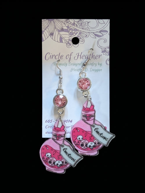 Pink and silver dangle earrings featuring cartoon potion bottles with skull details and a banner that reads 'To Auctions,' displayed on a branded backing card against a black background.
