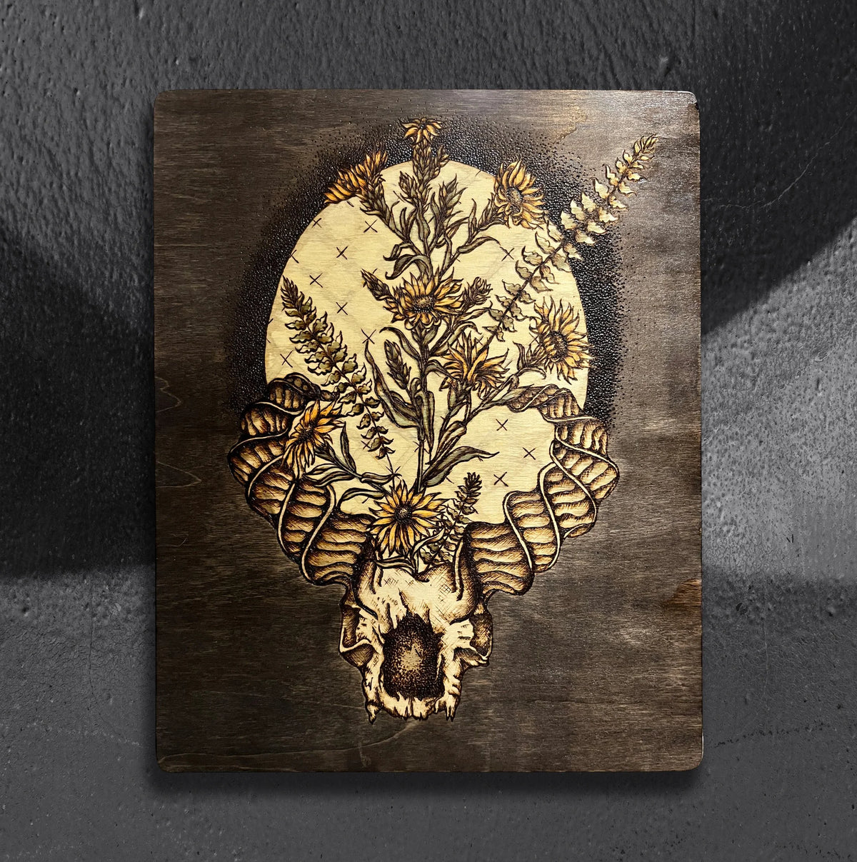Flora Capricora by CamiThunderInk, an original pyrography, watercolor, and stain painting on pine of a horned goat skull bursting into sunflowers and botanicals in front of a moon, on a stippled black background