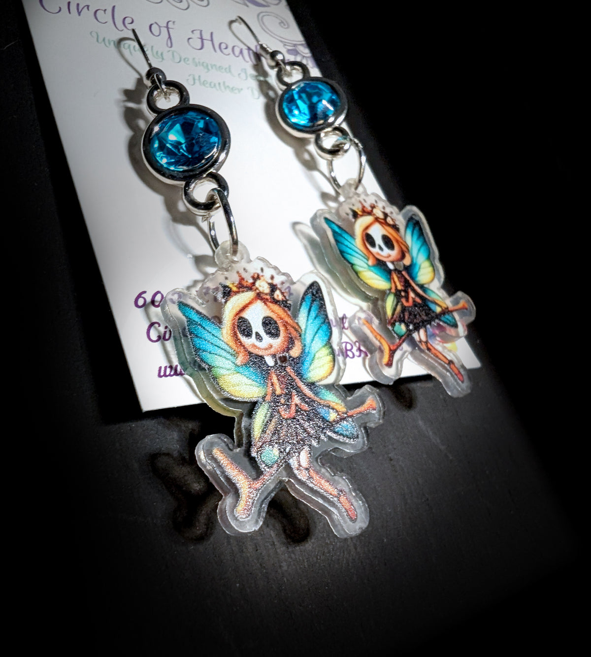 A pair of dangling fairy earrings displayed on a branded card against a dark background. Each earring has a blue faceted gemstone and a clear acrylic charm shaped like a fairy with multicolored wings and a skeleton-like face.
