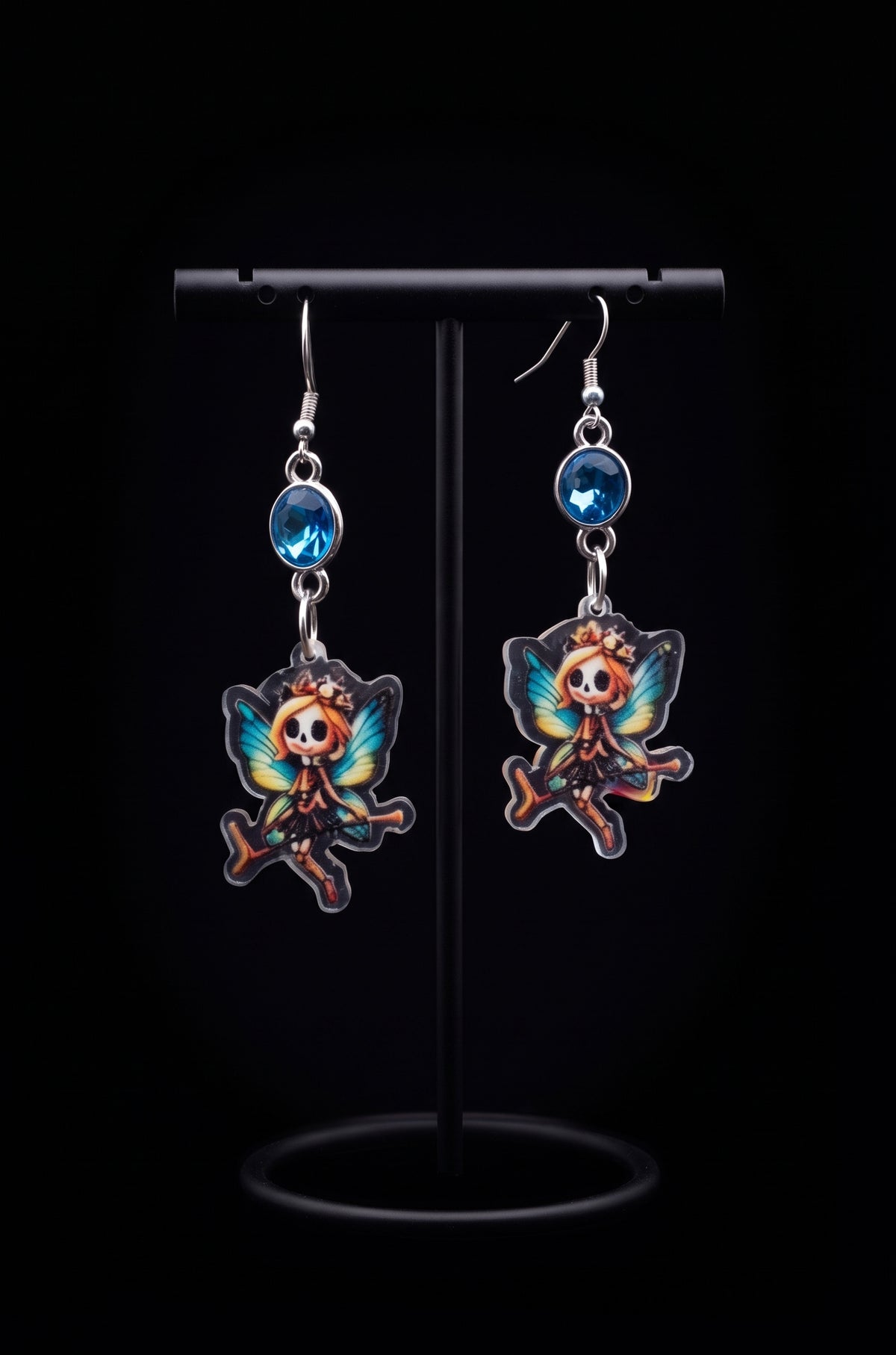 A pair of dangling earrings displayed on a black stand against a black background. Each earring has a silver fishhook wire, a round blue bead, and a colorful butterfly-shaped charm featuring a cartoon fairy design with orange, blue, and black details.