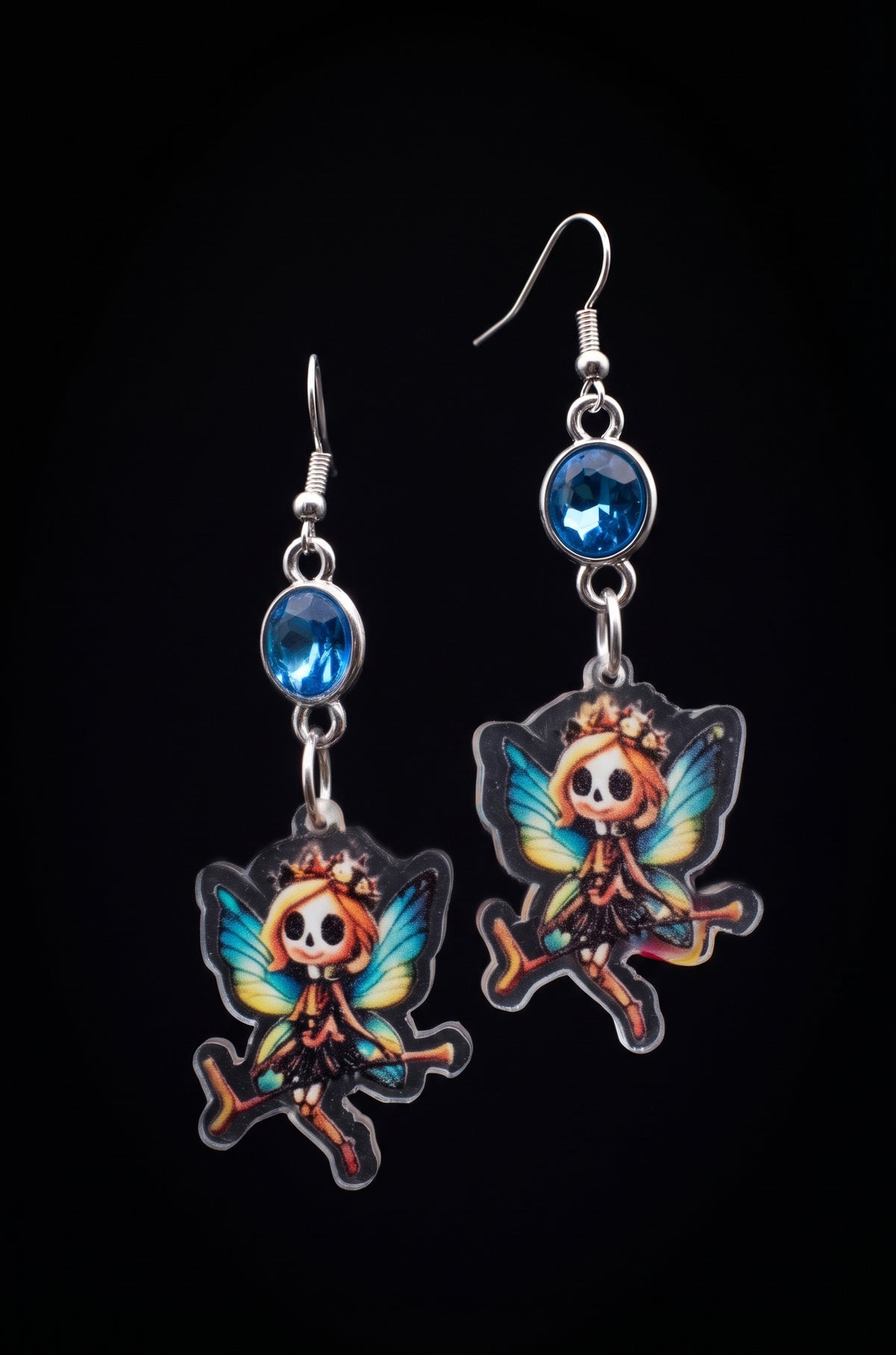 A pair of gothic fairy drop earrings featuring cartoon-style fairy charms with colorful wings, silver hooks, and round blue gemstones, set against a solid black background.