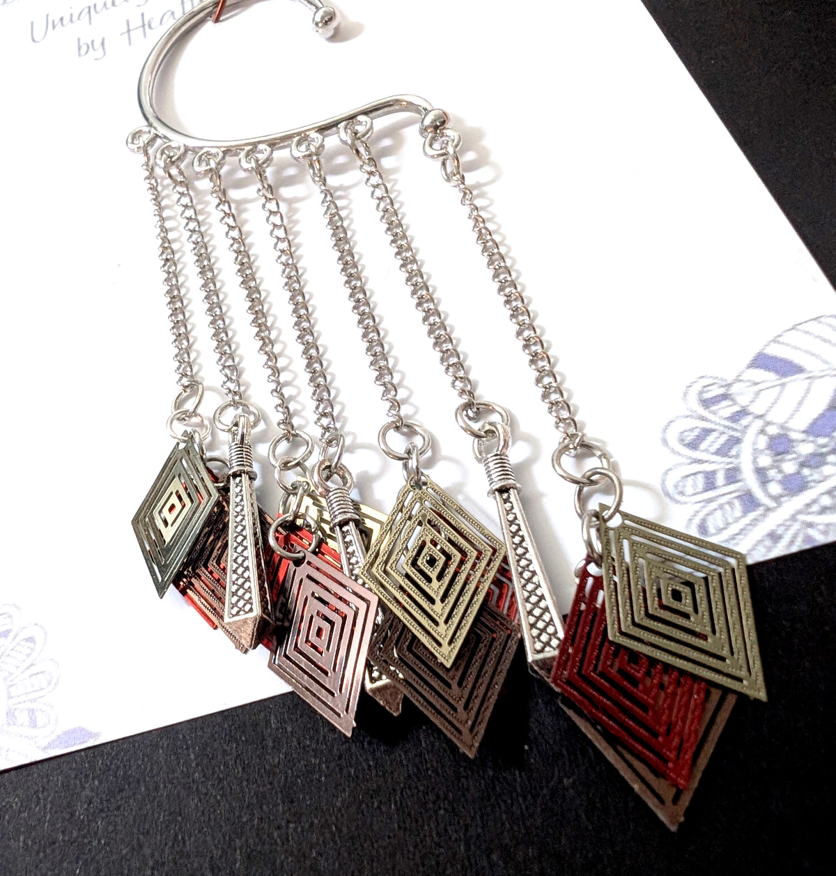 Close-up view of the intricate charms on the silver ear cuff, clearly showing the layered diamond construction (bronze, red, gold) and the textured silver rods against the backing card.