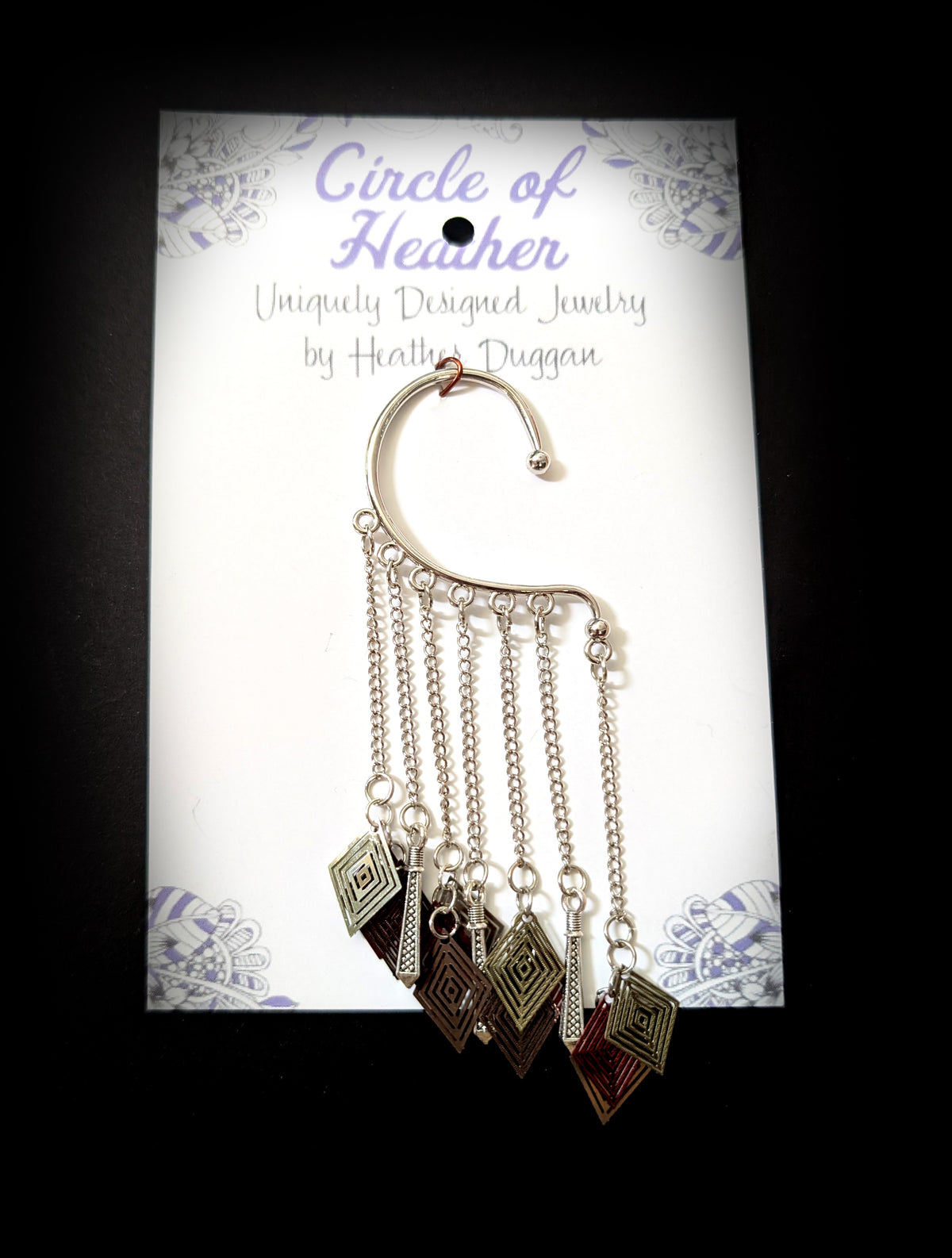 The silver geometric ear cuff attached to a white backing card featuring a decorative purple mandala pattern and text that reads "Circle of Heather, Uniquely Designed Jewelry by Heather Duggan."