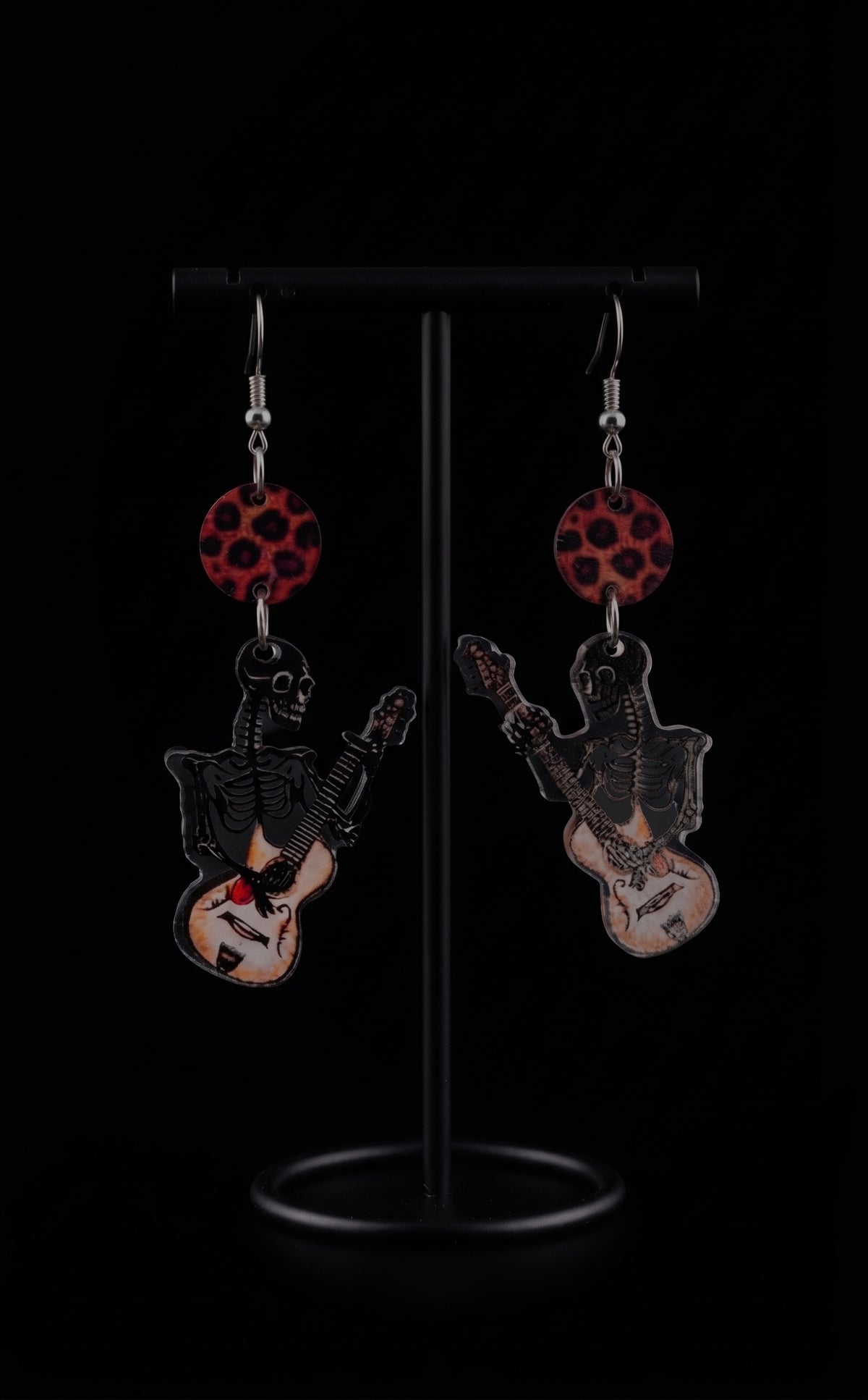 Guitar Ghoul Earrings by Circle of Heather, red and black leopard print discs above printed skeleton and guitar charms, hanging on a black T-bar jewelry stand against a black background.