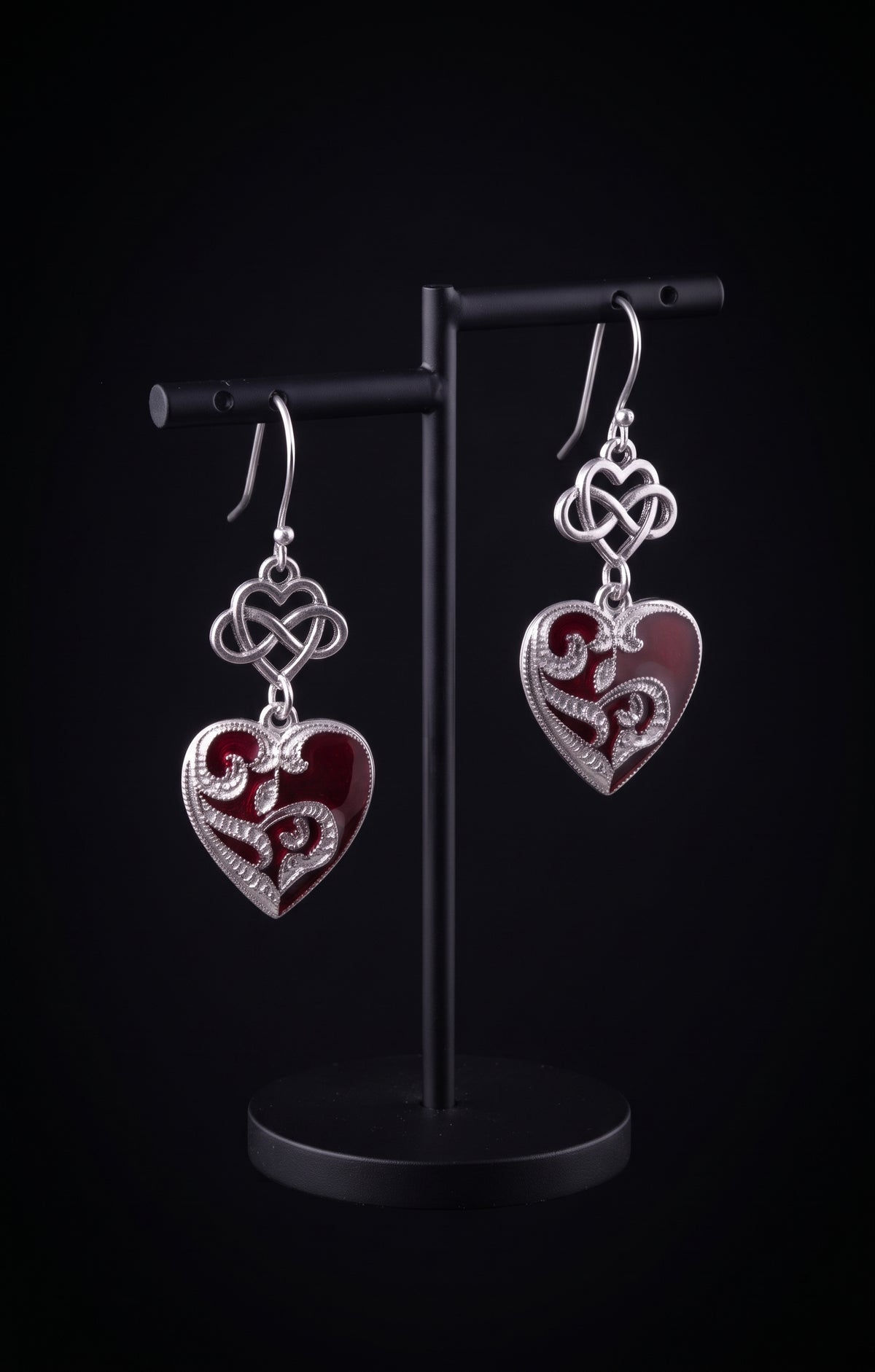 Infinity Hearts Earrings by Circle of Heather displayed on a black jewelry stand, silver-tone dangle earrings with an infinity heart knot above a filigree heart in deep red enamel