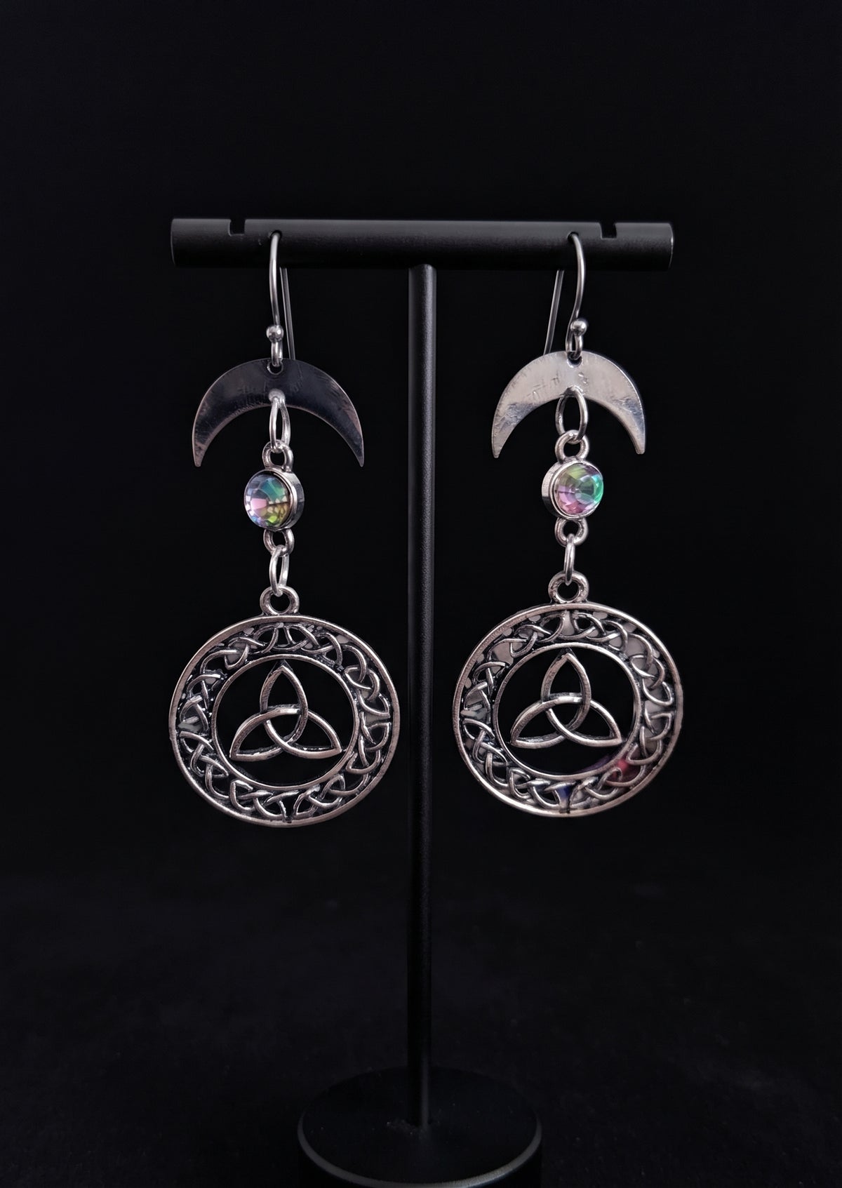 Knotted Luna Trinity Earrings by Circle of Heather, crescent moon tops with iridescent stones and round Celtic knotwork medallions, hanging on a black T-bar jewelry stand against a black background.