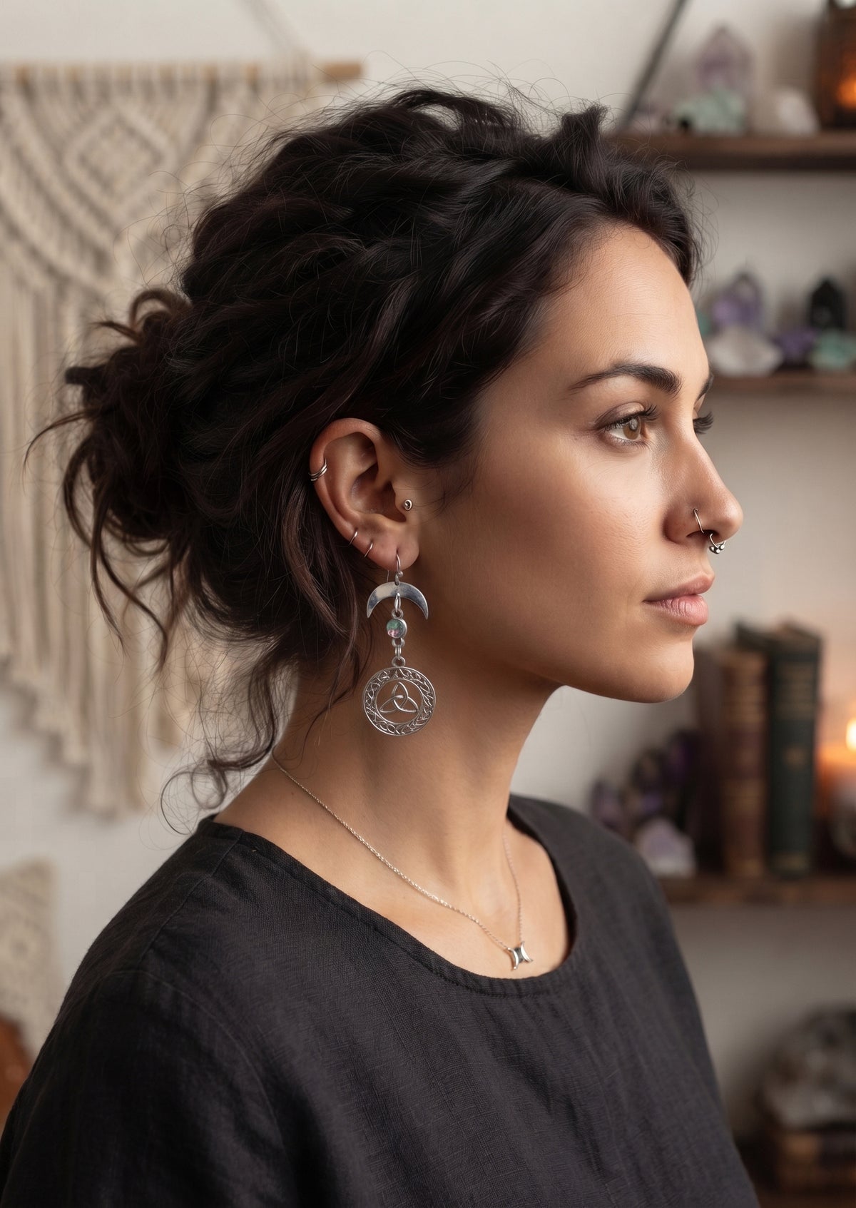 Knotted Luna Trinity Earrings by Circle of Heather worn by a model with dark hair in a loose updo, silver crescent moon above an iridescent stone and a Celtic knotwork medallion with a trinity knot, shown with a black linen top in a room with crystals and macrame.