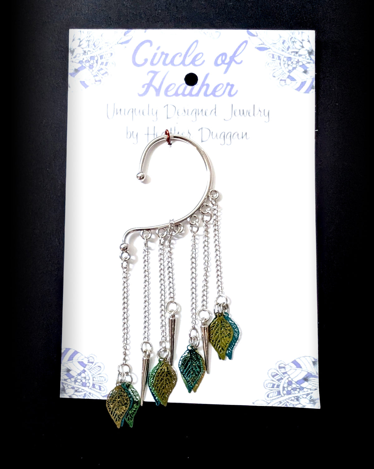 A silver leaf ear cuff attached to a white backing card featuring a decorative purple mandala pattern and text that reads Circle of Heather, Uniquely Designed Jewelry by Heather Duggan.