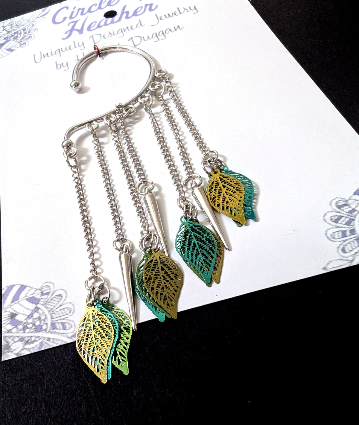 Close-up view of the silver leaf ear cuff showcasing the hanging chains, polished silver spikes, and layered green and gold filigree leaf charms.
