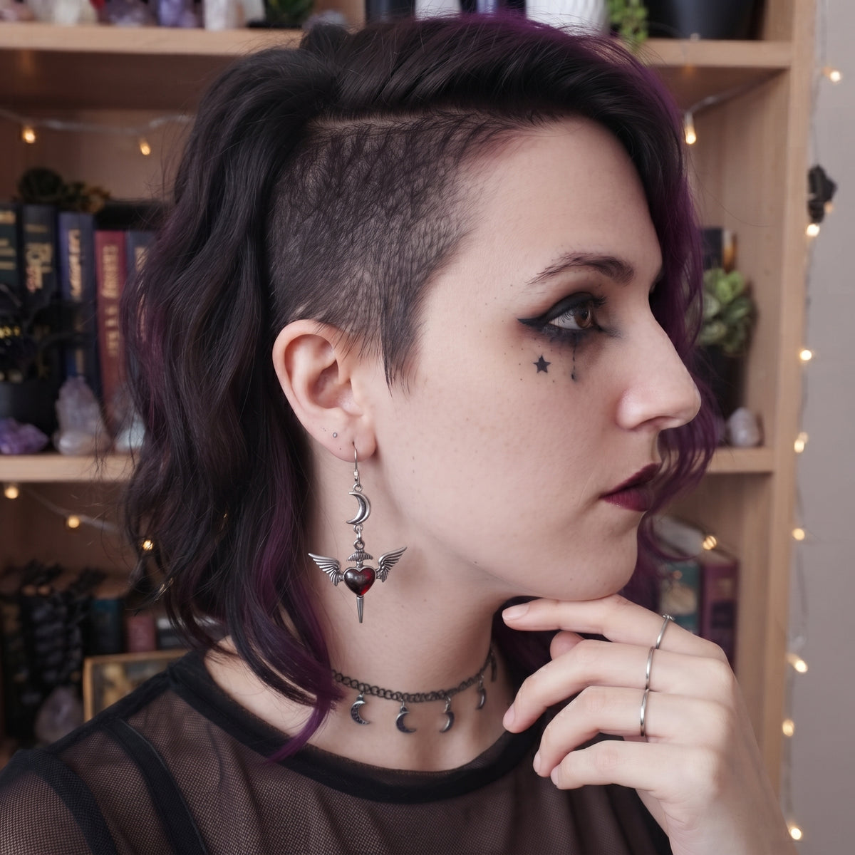 Love Soars Earrings by Circle of Heather worn by a model with dark purple hair, showing the crescent moon and winged heart dagger charm dangling near the ear