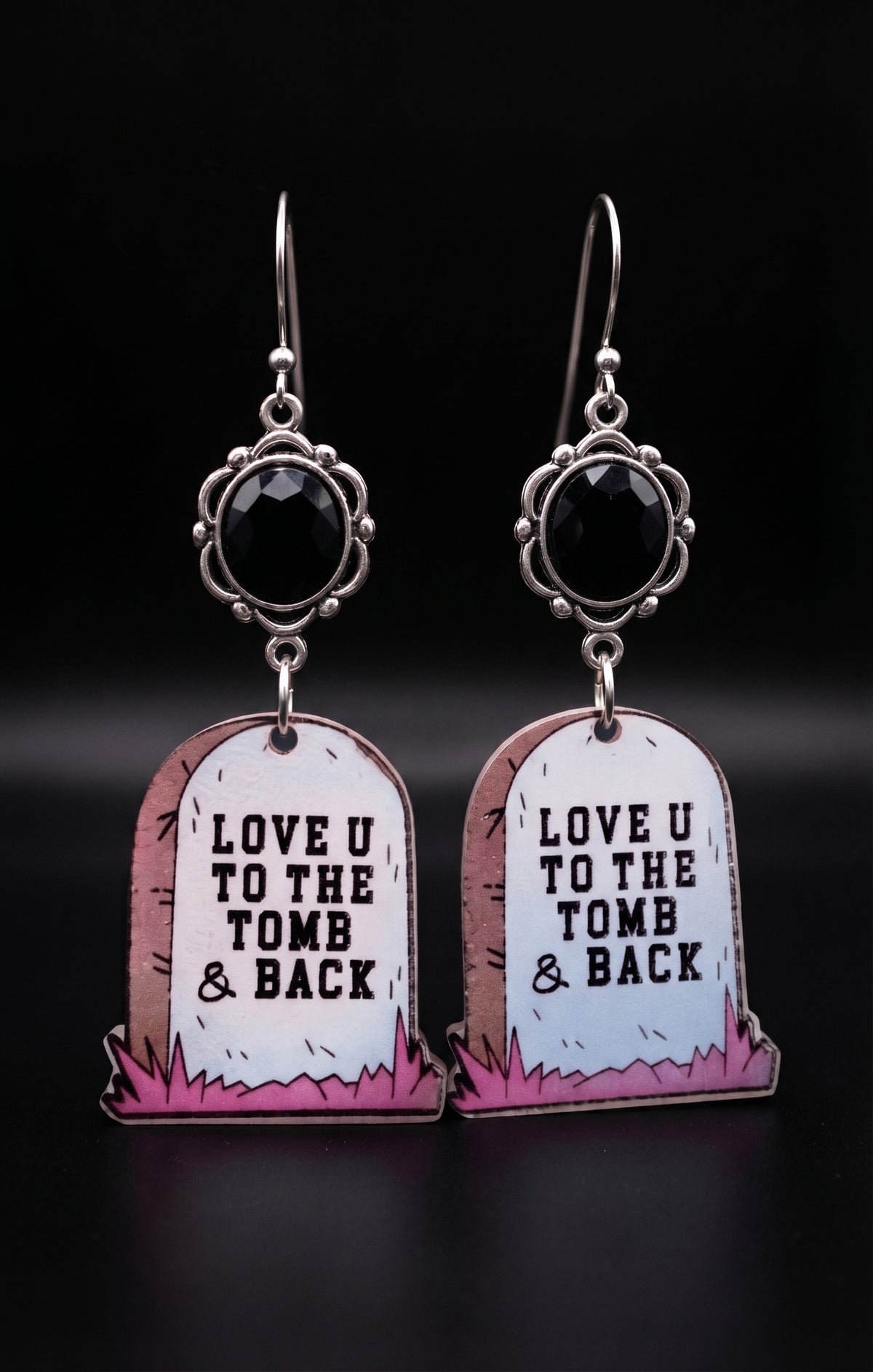 Love U to the Tomb and Back Earrings by Circle of Heather worn by a model with bright pink hair and wire glasses, faceted black stone above a printed headstone charm reading "Love U to the tomb and back."