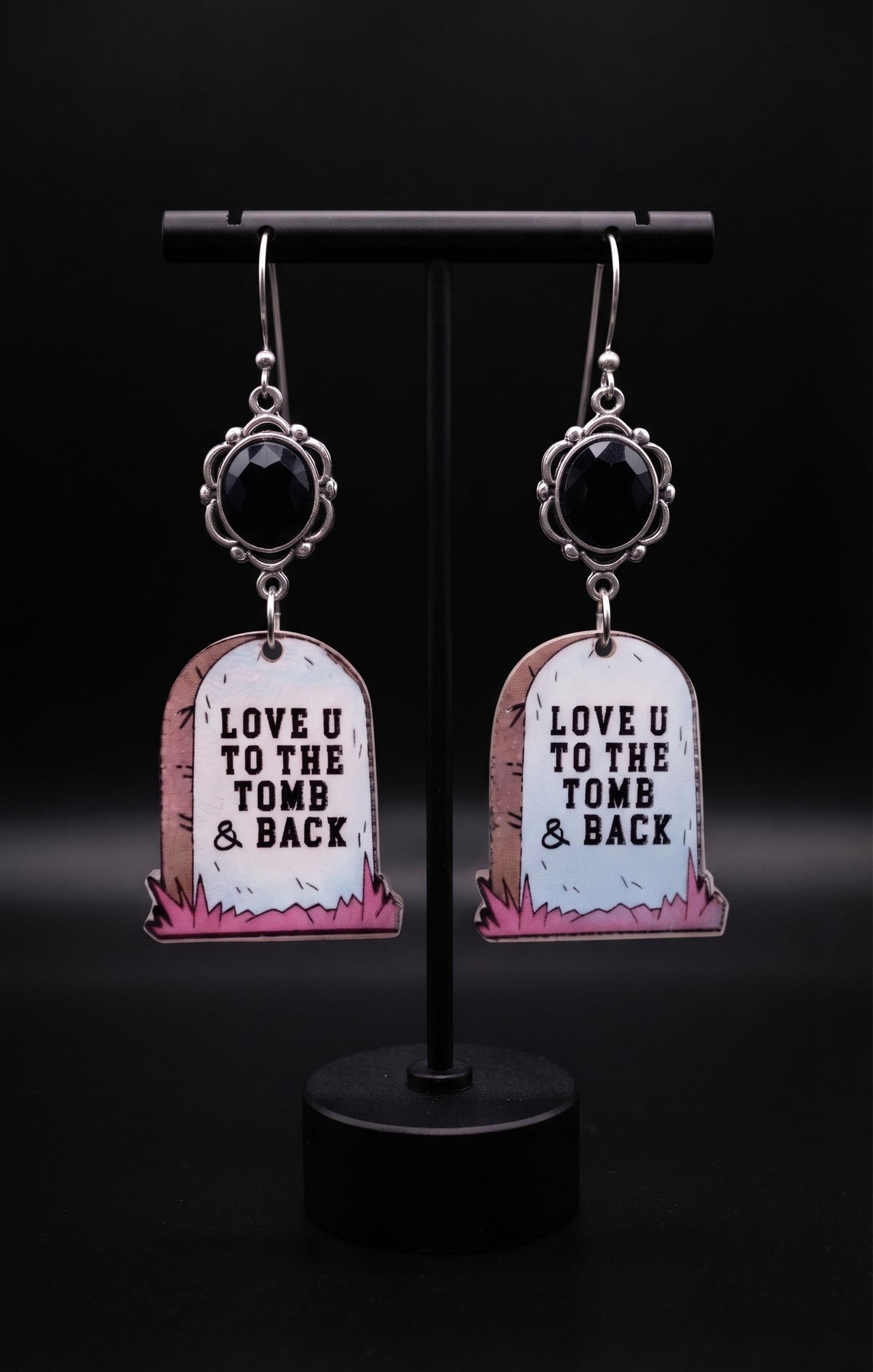 Love U to the Tomb and Back Earrings by Circle of Heather, faceted black stones in scalloped silver-tone settings above printed acrylic headstone charms, hanging on a black T-bar jewelry stand.