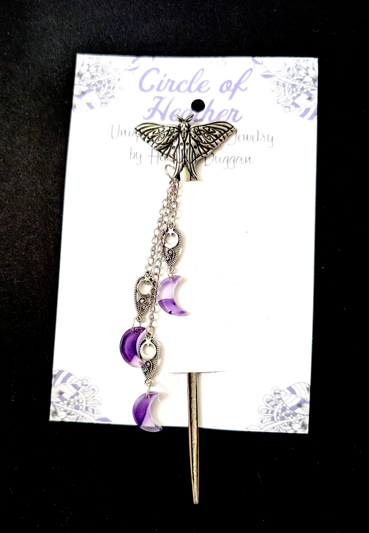 A silver lunate hair stick displayed on a white branded backing card reading "Circle of Heather Unique Jewelry by Heather Duggan." The piece features a detailed silver moth at the top with dangling silver chains, filigree charms, and translucent purple crescent moon beads.