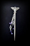 A full-length vertical shot of the silver moth hair stick against a dark textured background, highlighting the entire pin and the cascading arrangement of silver chains and purple crescent moon charms.