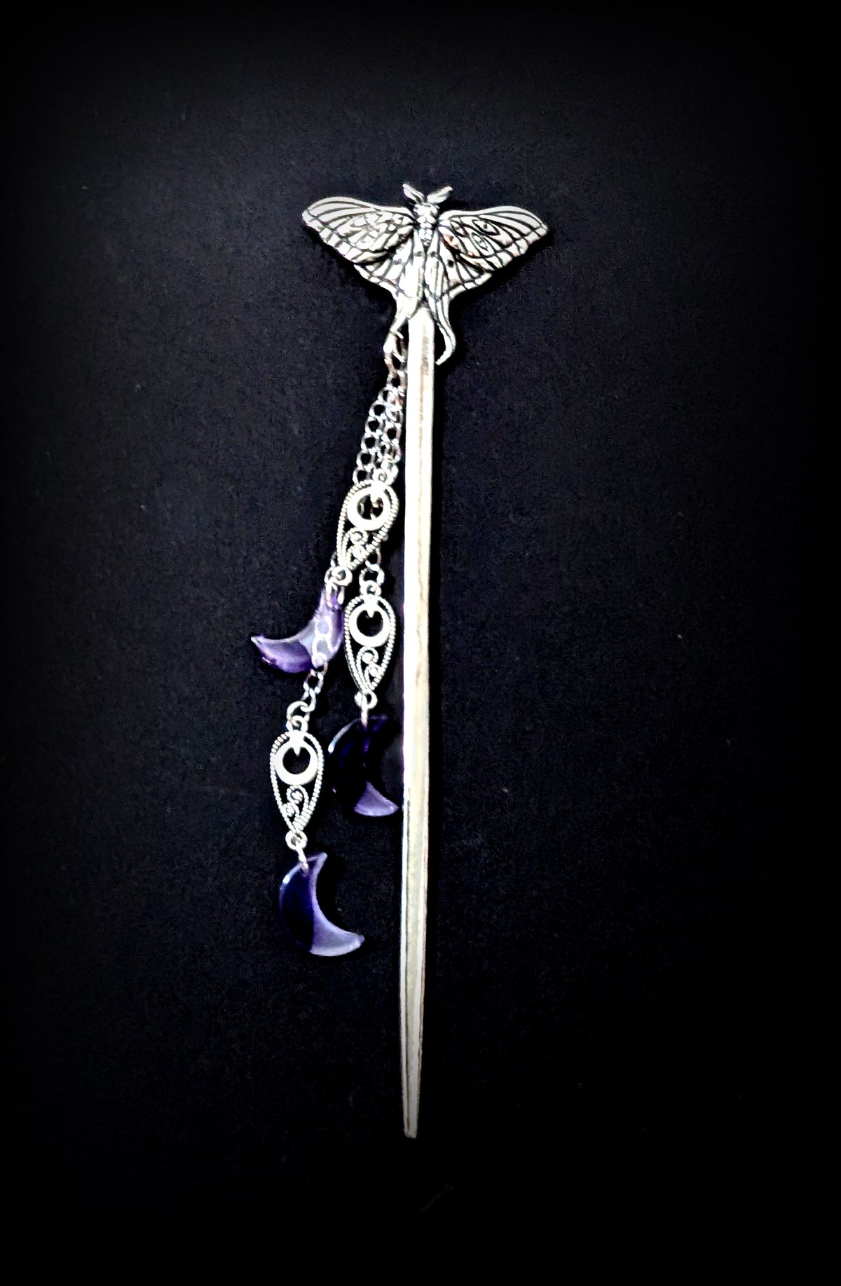 A full-length vertical shot of the silver moth hair stick against a dark textured background, highlighting the entire pin and the cascading arrangement of silver chains and purple crescent moon charms.
