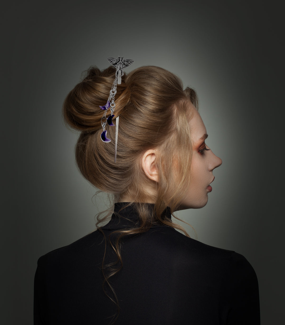 A profile shot of a person with blonde hair styled into an updo, wearing a black turtleneck against a dark gradient background. The silver moth hair stick from the previous shots is secured in the hair, with its dangling chains and purple crescent moon charms cascading down the side of the bun.