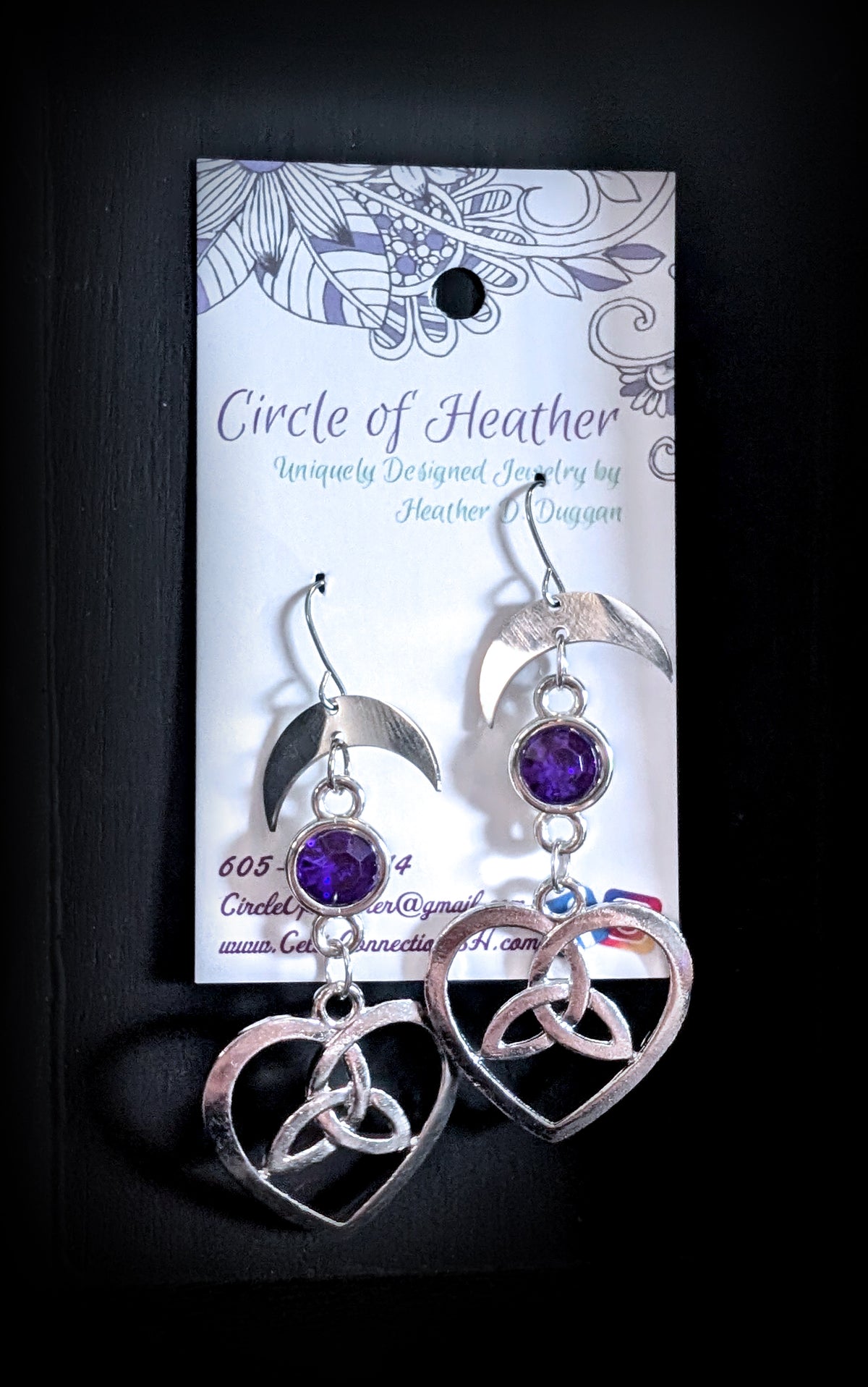 Lunar Love Trinity Earrings by Circle of Heather, crescent moon tops with purple stones and Celtic trinity heart charms, hanging on a black T-bar jewelry stand against a black background.