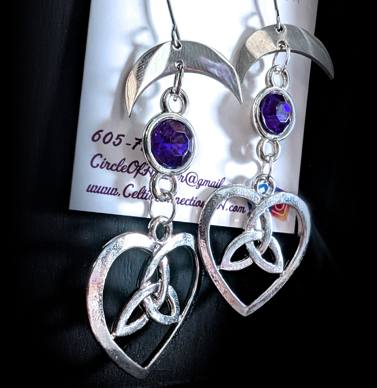 Lunar Love Trinity Earrings by Circle of Heather, angled close-up showing the faceted purple stone in its silver bezel and the trinity knot inside the heart charm, shown on the branded Circle of Heather card.
