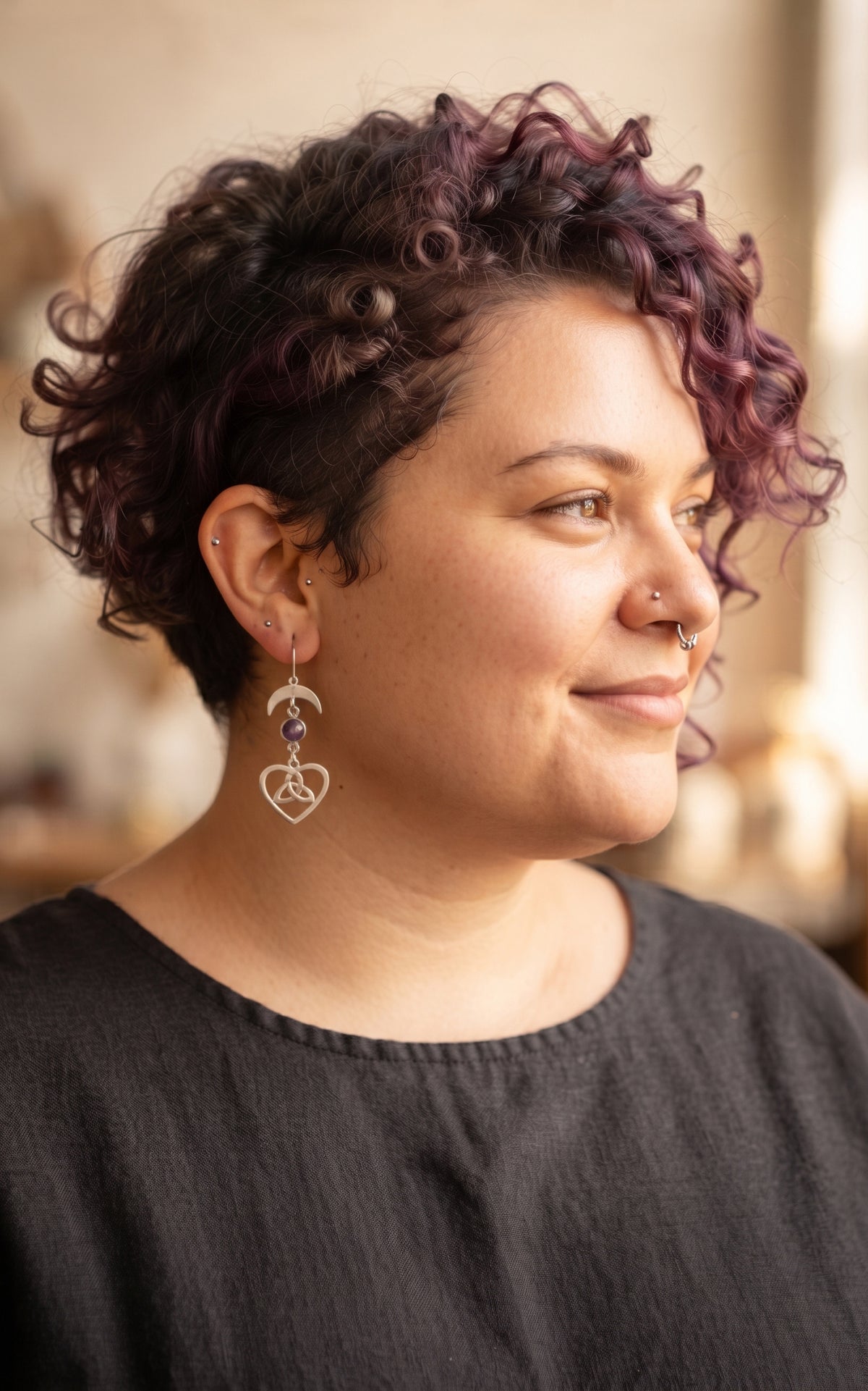 Lunar Love Trinity Earrings by Circle of Heather worn by a model with short curly dark hair with purple, silver crescent moon above a purple stone and a Celtic trinity heart charm, shown with a charcoal linen top.