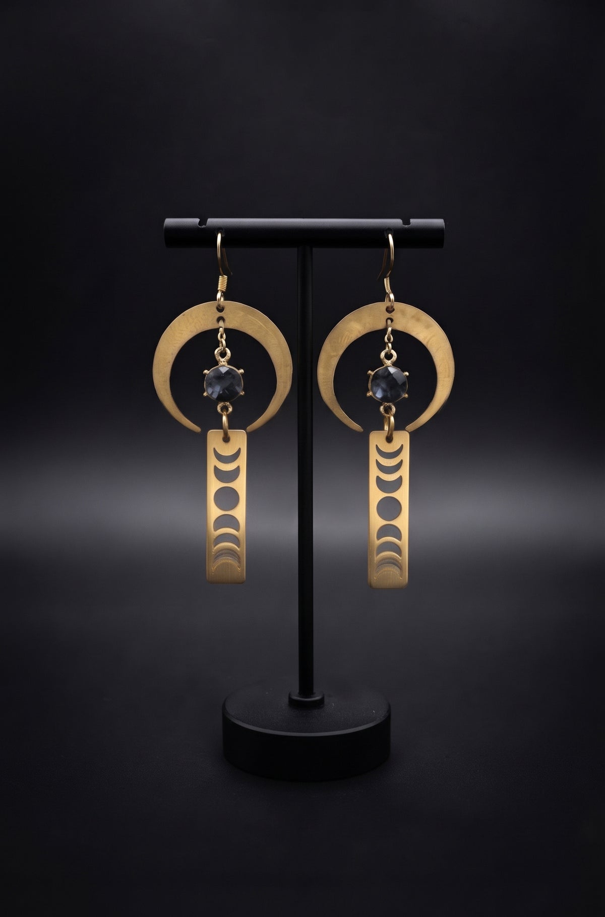 Lunar Obelisk Earrings by Circle of Heather, brushed gold crescent rings with grey blue faceted stones above long cut moon phase bars, hanging on a black T-bar jewelry stand.
