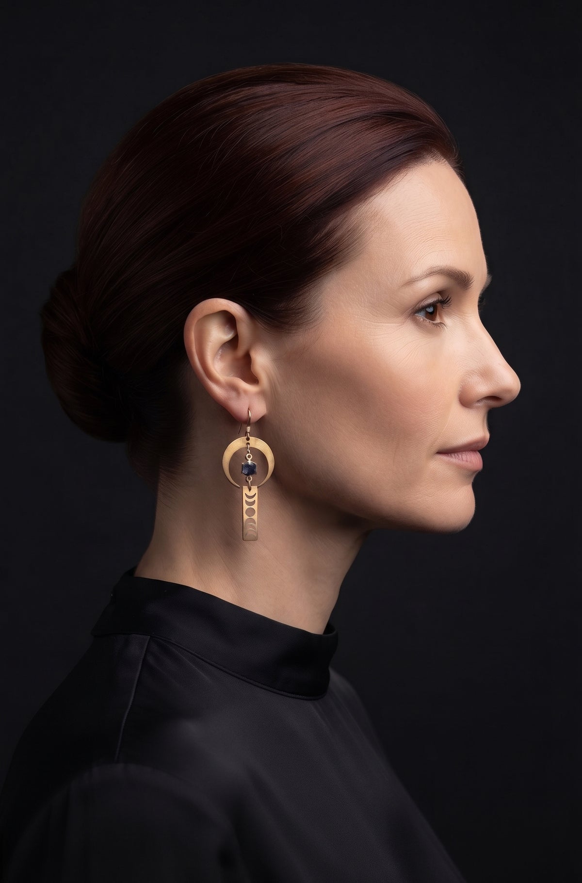 Lunar Obelisk Earrings by Circle of Heather worn by a model with auburn hair in a low bun, gold crescent ring with a grey blue stone above a long gold bar with moon phases cut through it, shown with a black mock neck top.