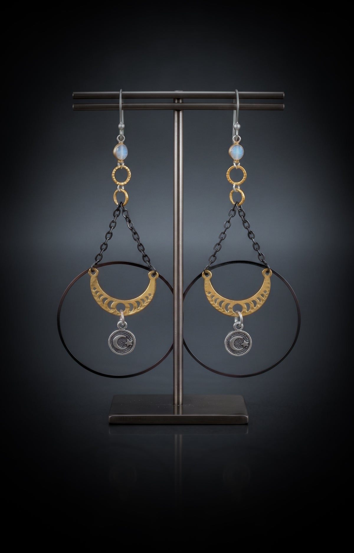 Chandelier Moonphase Earrings by Circle of Heather, both earrings hanging on a dark metal T-bar jewelry stand, showing the full length from the opalescent stone down to the silver moon and star medallion.